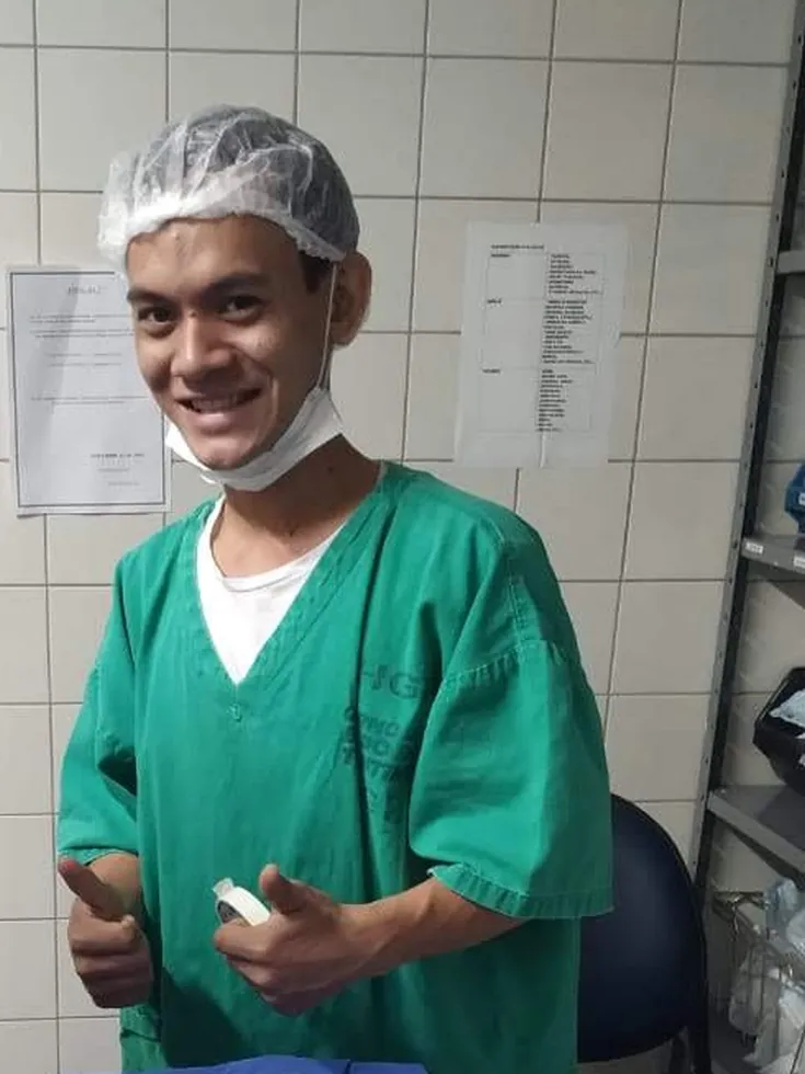 Thalles Diamantino de roupa privativa verde e touca, sorrindo ao lado de um pacote de material hospitalar