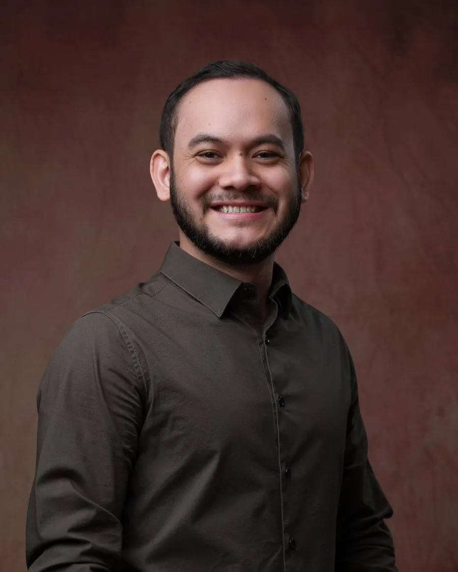 Thalles Diamantino em pé, sorrindo, de camisa escura sobre fundo marrom